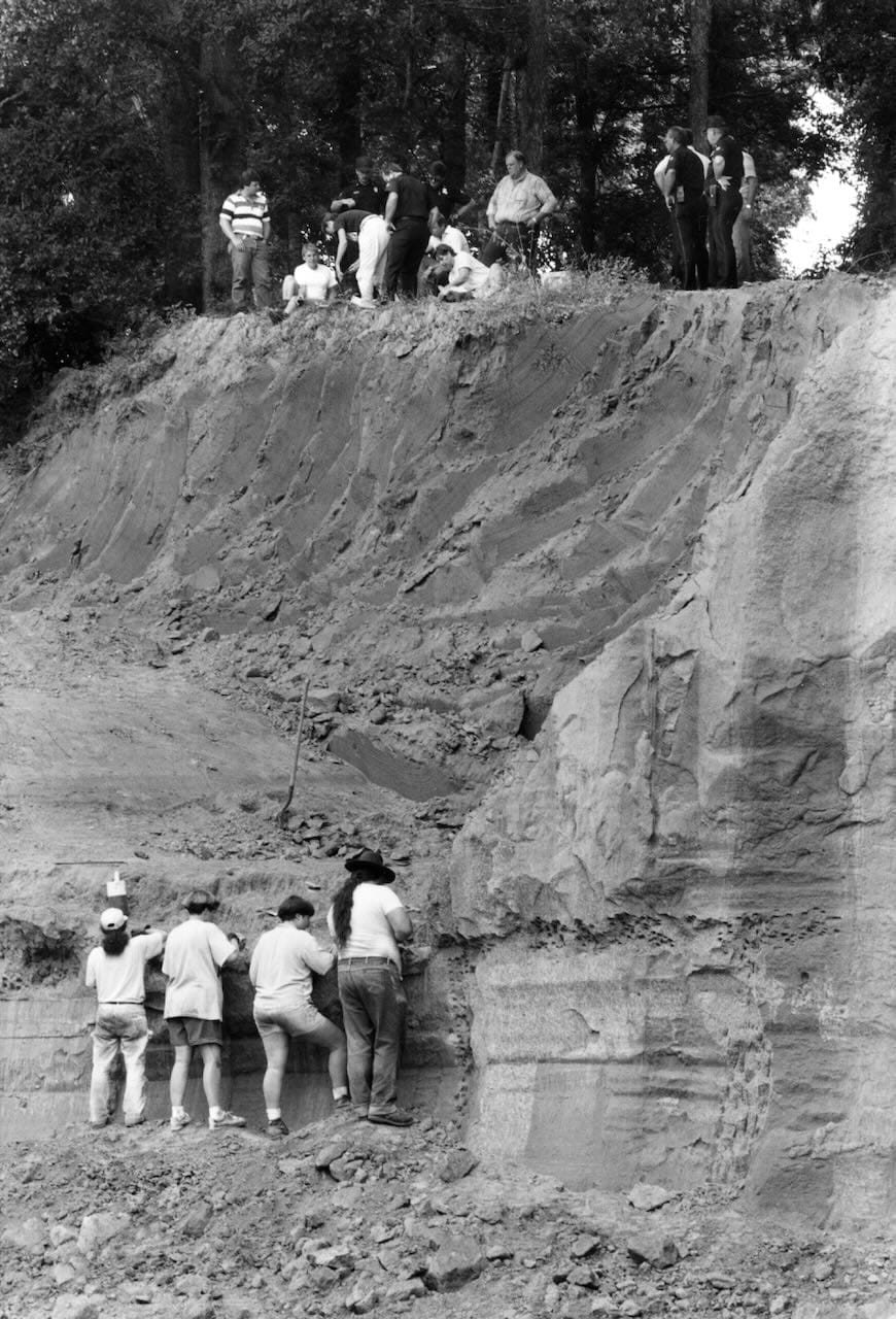 In a black and white photo, 4 people dig at the bottom of a 20-foot-tall cliff. At the top of the cliff, another group of scientists & prison staff work on a 2nd excavation.
